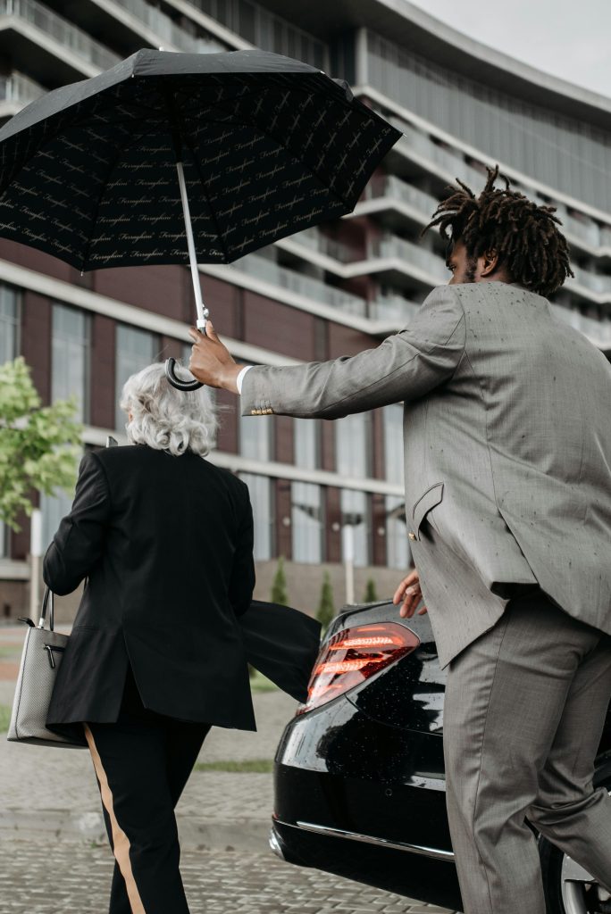 Two business professionals with an umbrella near a luxury car in an urban setting.